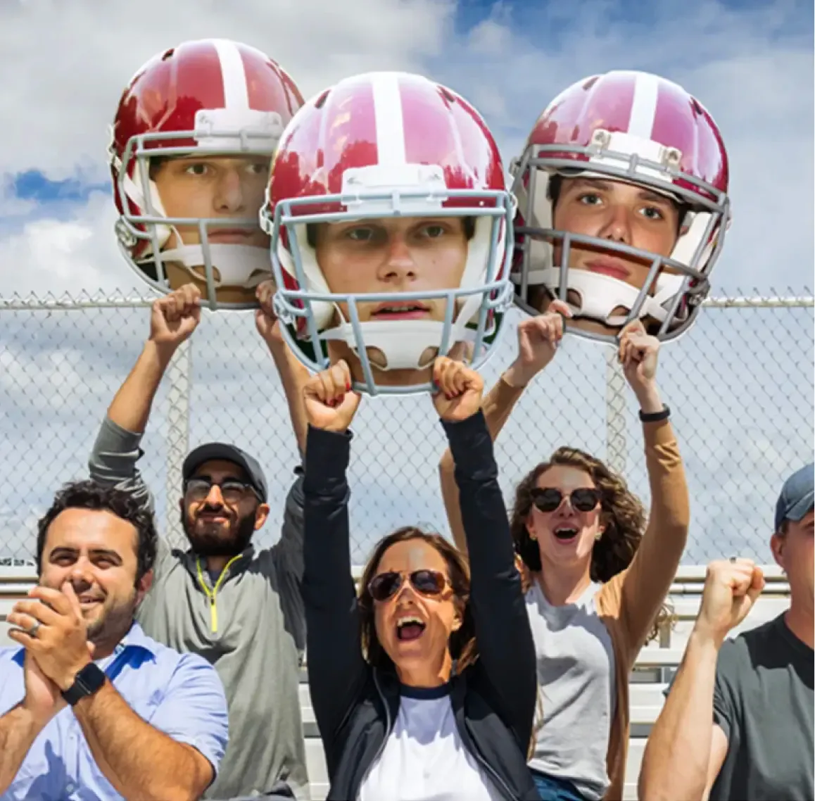 Group of fans cheering at a sports event with three holding large cutouts of football players' heads wearing red helmets.